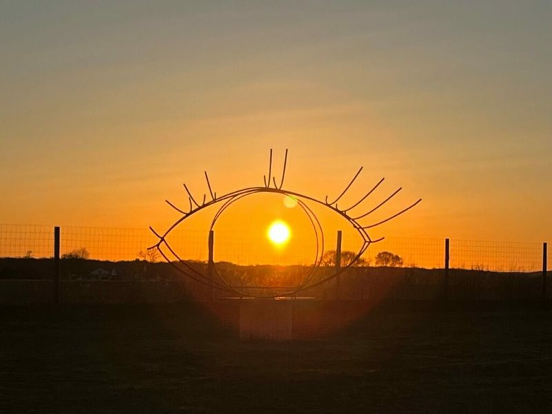 A thin steel outline of an eye with lashes in a field, with the setting sun in the middle of it