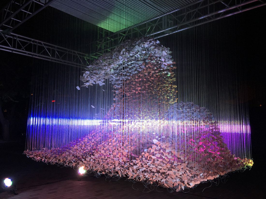 A wave of bricks hanging on thin lines under a steel truss, lit with purple and blue light at night