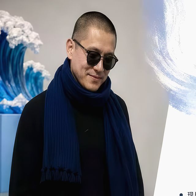 Mr. Zhang in sunglasses and a navy scarf, standing beside a sculpture of a blue wave